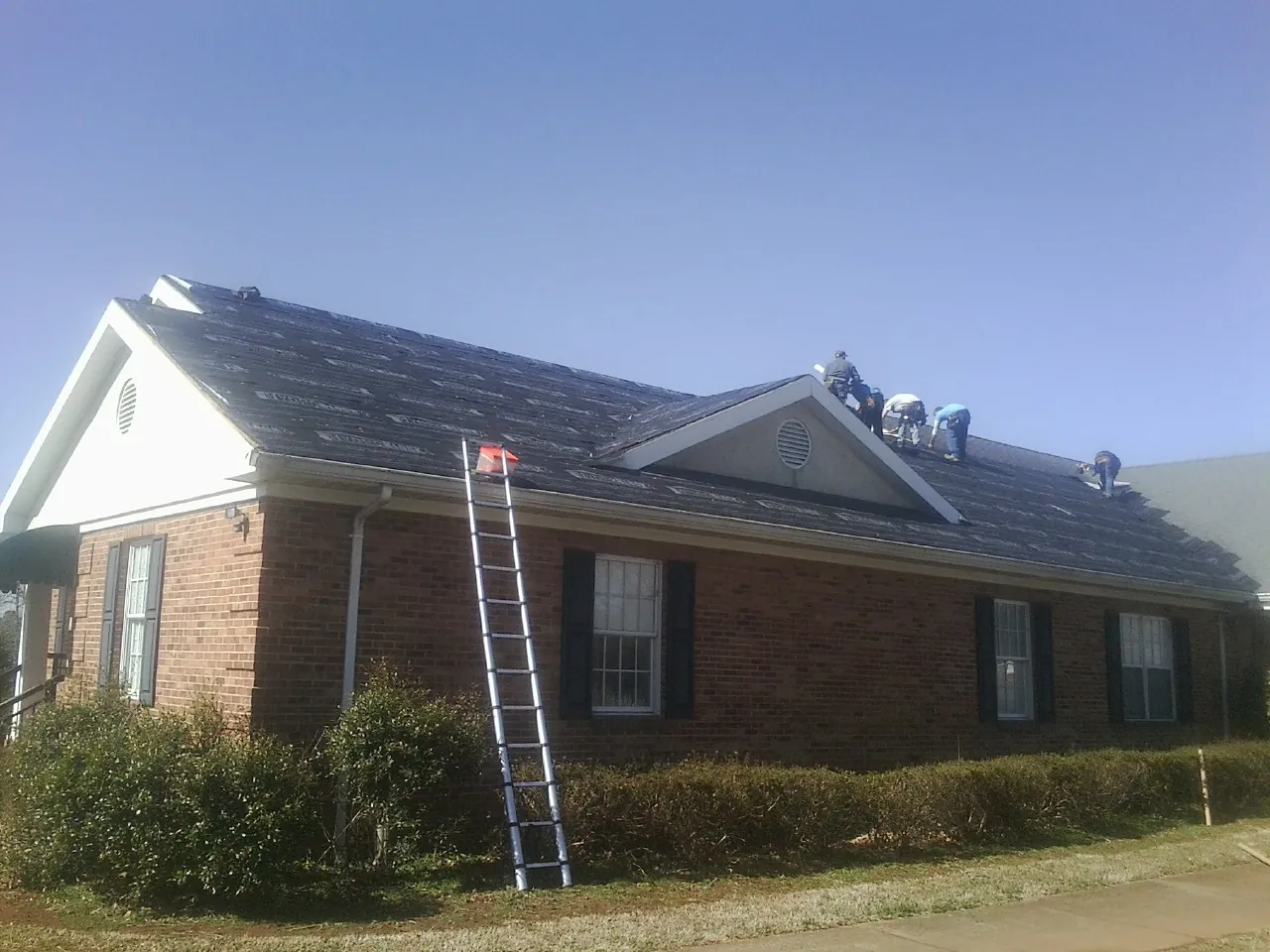 Crew tearing shingles off a brick ranch roof with a ladder set — Emergency Roof Repair work by our crew in Saint Rose