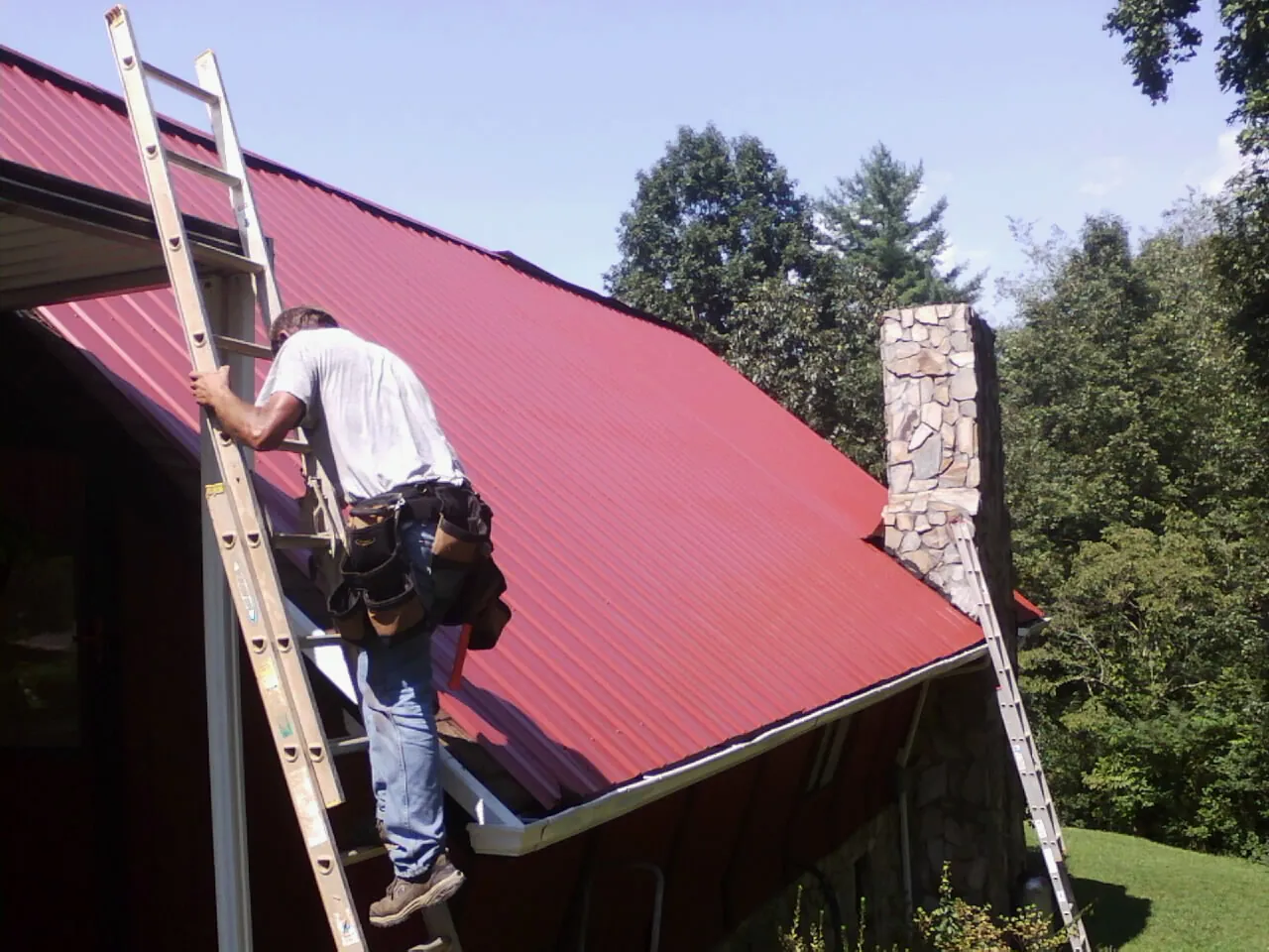 Roofer on a ladder beside a red metal roof and stone chimney — Soffit & Fascia Repair by Roofing Brothers in Saint Rose